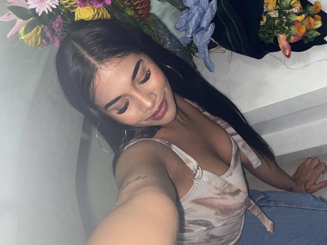 IsisFerrer with long black hair, wearing a tie-dye tank top, hoop earrings, and a nose ring, taking a selfie, showing her tits and a tattoo on her arm.
