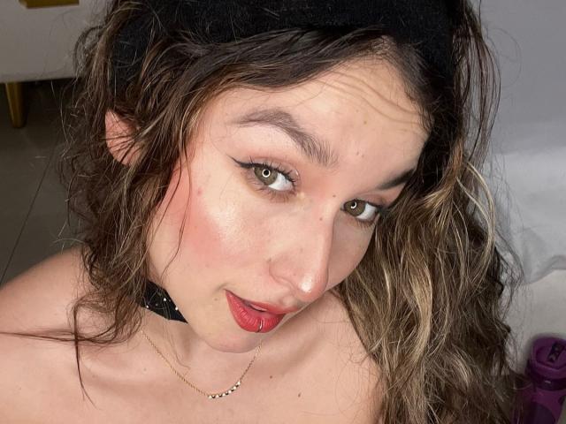 MilaGonz with wavy brown hair, green eyes, black headscarf, red lipstick, and a black choker necklace, looking up at the camera with a slight smile. Her boobs are visible.