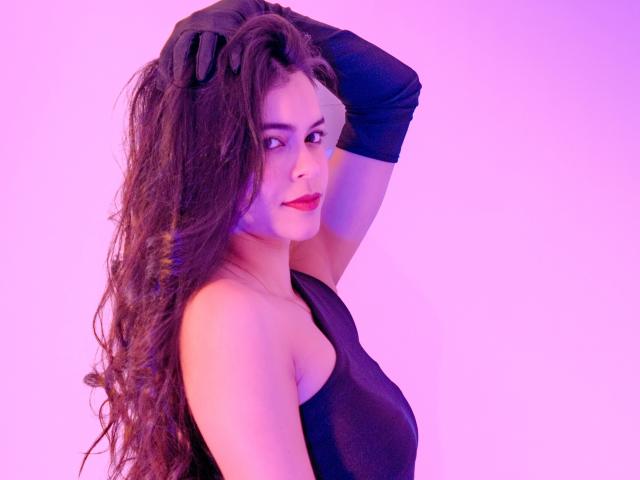AlanaBlake with long curly brown hair, black sleeveless top, and black gloves, raising her hand to her head, smiling with red lipstick, against a pink-purple background.