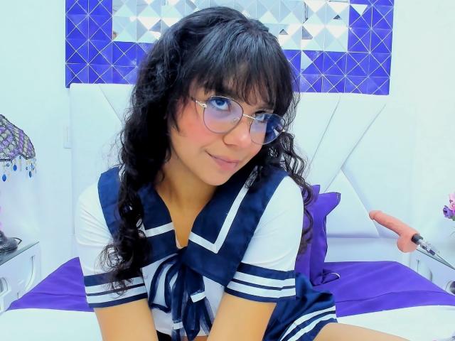 Julia Bennett, con cabello negro rizado, gafas y un uniforme escolar azul marino y blanco, sentada en una cama con sábanas moradas, sonriendo, mostrando sus pequeños senos y su linda cara..