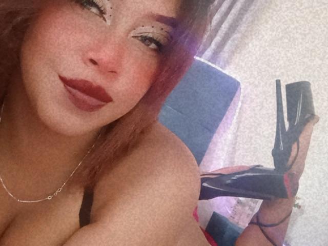 Dalila Heinz, com batom vermelho, delineador marcante e cabelo com tons de roxo, segurando sapatos de salto alto pretos, mostrando os seios, usando um sutiã preto e um colar de prata..