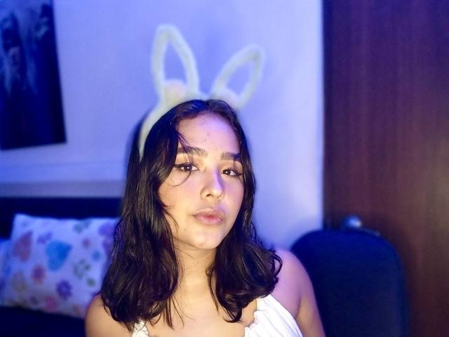 AbyGailgomez, avec ses cheveux noirs et ondulés, portant un haut blanc à une épaule et un serre-tête jaune en forme de lapin duveteux, regarde l'appareil photo avec une légère moue..