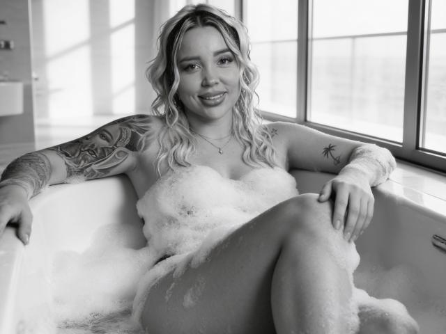 MiaBlondeHot in a bathtub, covered in bubbles, showing her tits, with tattoos on her arms, smiling, legs crossed, blonde wavy hair, black and white photo.