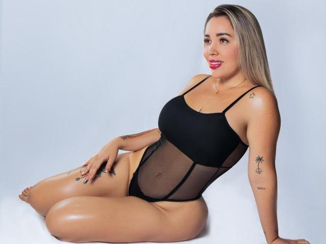 MiaBlondeHot sitting on floor, wearing black sheer bodysuit, showing her curvy ass and tits, blonde hair, tattoos on arms, smiling with red lipstick, white background.