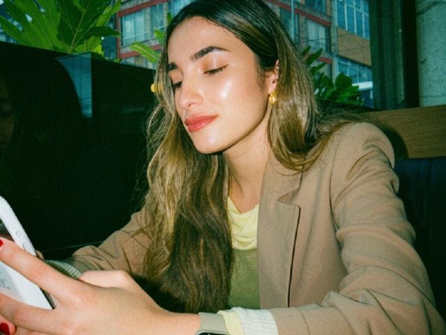 AdharaCooperr with long brown hair, closed eyes, and pink lipstick, wearing a tan blazer, holding a white tablet, sitting in a dark booth, smiling slightly.