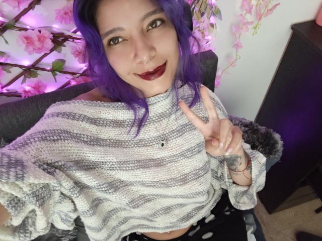 DarckVelvet, con cabello morado, aro en la nariz y lápiz labial oscuro, vestida con un suéter a rayas, mostrando un signo de la paz y revelando un tatuaje en su mano derecha. No se ven los senos..