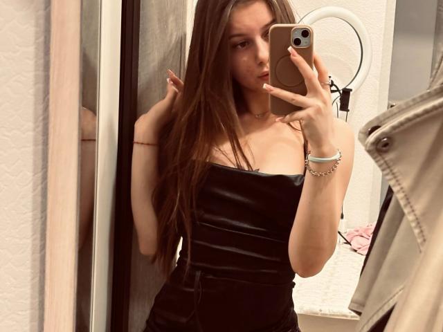 Kimberly Zampieri, met lang bruin haar, draagt een strak zwart leren jurkje en maakt een selfie in de spiegel met een gouden iPhone. Haar kleine borsten zijn zichtbaar onder de jurk..
