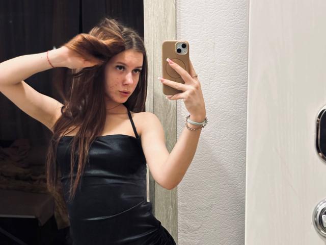 Kimberly Zampieri, con cabello largo y castaño, vestida con un vestido negro sin tirantes, sosteniendo un teléfono dorado, tomando una selfie en el espejo, con la mano en el cabello, mostrando sus pechos..