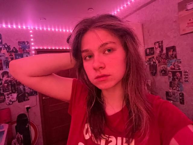 ShiraWolny with wet brown hair, red shirt, hand in hair, pink-lit room, photos on wall, serious expression, visible nose ring.