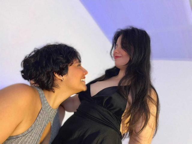 GabyDani with short black hair, gray top, leaning towards GabyDani with long black hair, large boobs, black dress, smiling. GabyDani with short hair looks up at GabyDani with long hair.