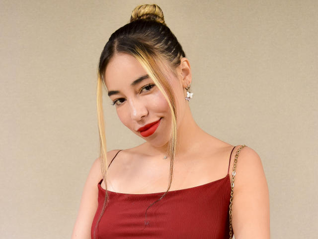 MissVallen, con cabello rubio recogido en un moño, vestido rojo, lápiz labial rojo y aretes con forma de estrella, sonriendo ligeramente, mostrando su trasero lindo y sus pechos pequeños. Está mirando a la cámara..