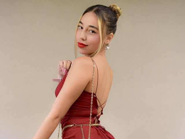 MissVallen com coque loiro, vestido vermelho, alça de corrente, batom vermelho, olhando por cima do ombro, segurando um anel rosa, costas nuas, brincos, sorrindo, pele clara, sexy, bonita..
