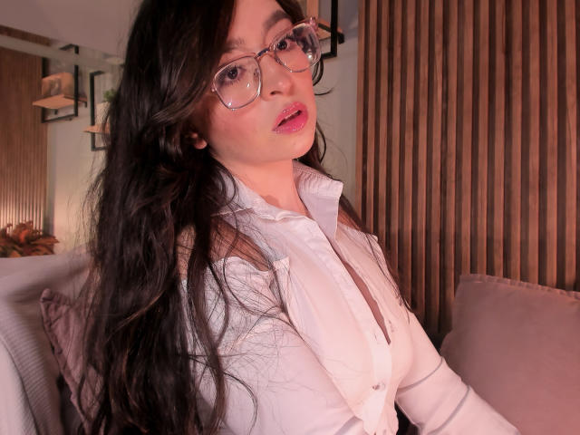 Eva McAllister, con lunghi capelli neri, occhiali, rossetto rosa e camicia bianca con bottoni, guarda la fotocamera, mostrando il seno, in un ambiente interno accogliente, con una parete di legno sullo sfondo..