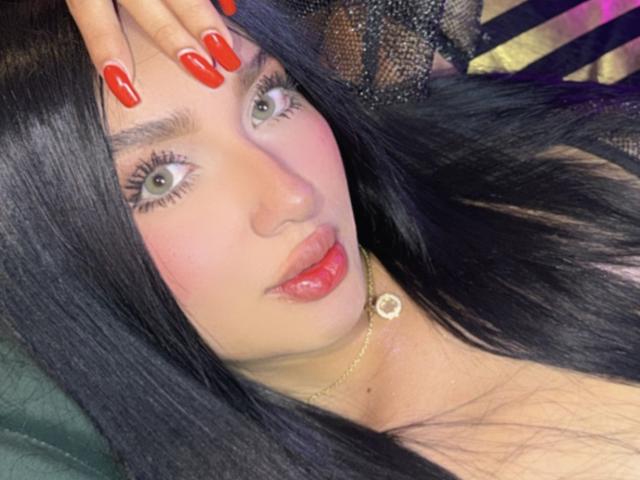 Lilly Taylor tem cabelos pretos longos, unhas vermelhas, olhos verdes, blush rosa e batom vermelho. Ela está usando um colar de ouro com um pequeno pingente e um padrão de renda..