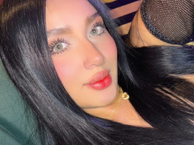 Lilly Taylor tem cabelos pretos longos, olhos verdes e lábios vermelhos, usando um colar de ouro, com uma manga de malha preta visível no lado direito. Seu rosto está levemente avermelhado..
