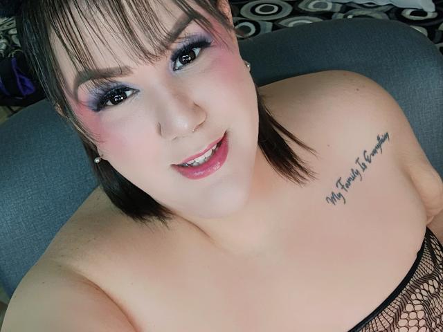 GiiannaMorrys with pink eyeshadow, purple lipstick, and a nose ring, smiling at the camera. She has black fishnet top, visible tattoo on right shoulder, and short brown hair.