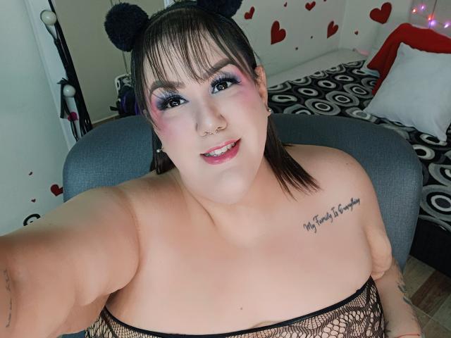 GiiannaMorrys with black fishnet top, cat ears, pink and purple eyeshadow, nose ring, tattoo on right chest, smiling, holding camera, showing large bare tits.