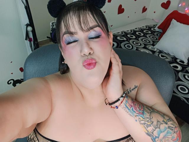 GiiannaMorrys with puffy pink lips, purple eyeshadow, black fishnet top, black hair buns, nose ring, tattooed arm, bracelet, sitting on gray chair, pouty expression.