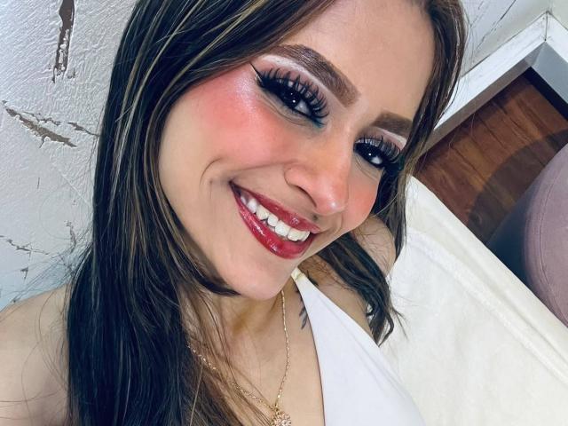 AfroditaWild with long brown hair, red lipstick, and big eyelashes, smiling at the camera, wearing a white halter top, showing off her cute nose and glowing skin.