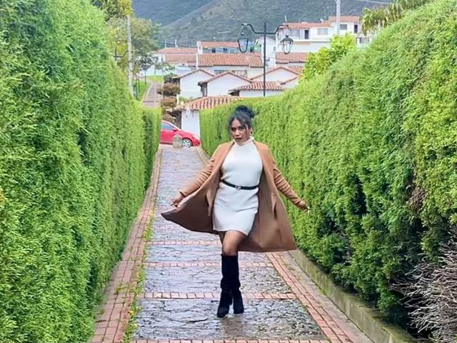 SophieSonyas, de figura voluptuosa con trasero grande, vestida con un vestido blanco, un abrigo marrón, botas negras, un cinturón negro, setos verdes, un camino de adoquines mojados, sosteniendo el abrigo abierto, de pie con confianza, calle mojada..