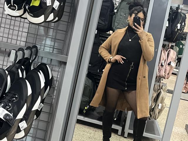 SophieSonyas con cabello corto y rizado, abrigo color piel, vestido negro, medias negras, sosteniendo un teléfono en el espejo, mostrando su trasero, en una tienda con mochilas y bolsos en los estantes..