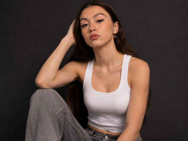 DanicaStuer with long brown hair, white tank top, small tits, gray pants, sitting, hand on head, looking serious, gold earrings, dark background, soft light, slight smile.