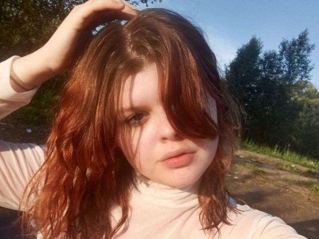 VeldaWesterbeck has red, wavy hair covering one eye, fair skin, and is wearing a white turtleneck, with a hand in her hair, in a sunny outdoor setting.