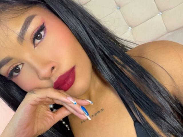 LexieBloom, con cabello negro largo, lápiz labial rojo, uñas azules y un tatuaje en el pecho, apoyando el mentón en la mano, mirando a la cámara..