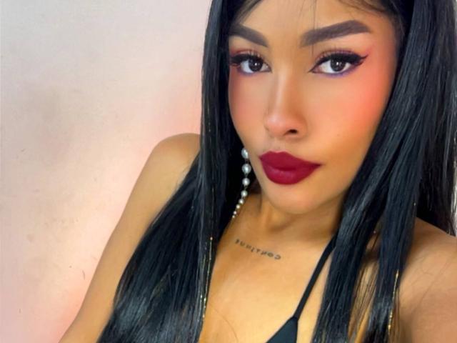 LexieBloom tem cabelos pretos longos, batom vermelho e delineador marcante. Ela está vestindo uma blusa preta e usa brincos de pérolas. Sua pele bronzeada tem uma tatuagem logo abaixo dos seios..