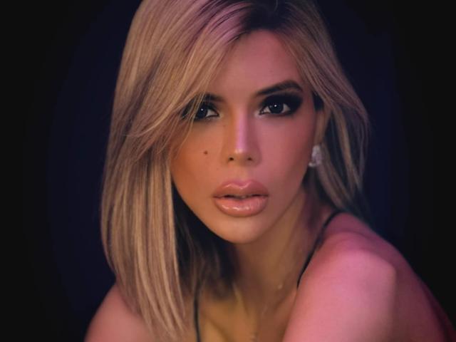 HotCumAdriana69x con capelli biondi, occhi scuri, labbra lucide e un neo sulla guancia sinistra, che indossa una collana e guarda seducentemente la fotocamera..