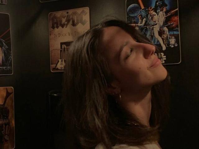 JennyMyLuv's head is tilted back with a small smile, eyes closed, brown hair, earrings, and a slightly flushed face in a dimly lit room with rock posters.