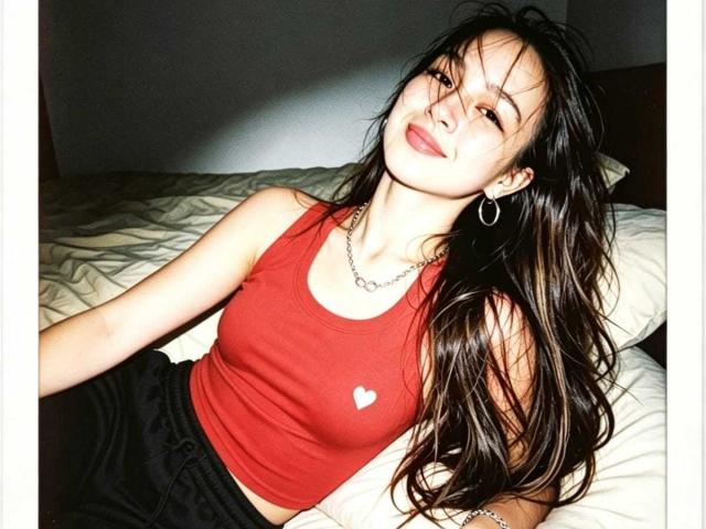 MilaHans con cabello largo y castaño, camiseta roja que muestra un pequeño corazón en el pecho, pantalones negros, collar de plata, aros grandes, sonriendo, guiñando un ojo, acostada en la cama con una almohada blanca..