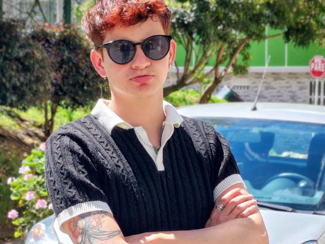 GabyThoms, con cabello rojo corto, gafas de sol negras, suéter negro con cuello blanco, brazos cruzados, tatuaje en el brazo derecho, anillo en la mano izquierda, de pie frente a un coche blanco..