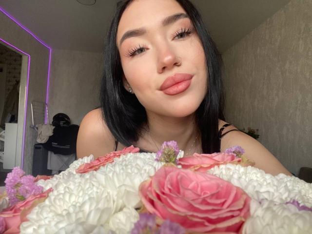 DiannLamphear, avec ses cheveux noirs, ses lèvres pleines roses, et ses longs cils, arborant une expression boudeuse, tient un bouquet de roses roses et de chrysanthèmes blancs au premier plan..