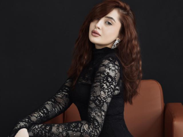 MilanaVesterli with wavy red hair, wearing a black lace dress, sitting on a brown leather chair, looking over her shoulder, showing her tits through the lace.