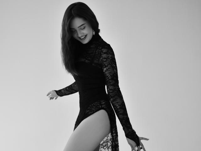 Black-and-white photo of MilanaVesterli in a lace long-sleeve dress, showing her thigh, smiling with her eyes closed, and her hand on her hip. She has medium tits.