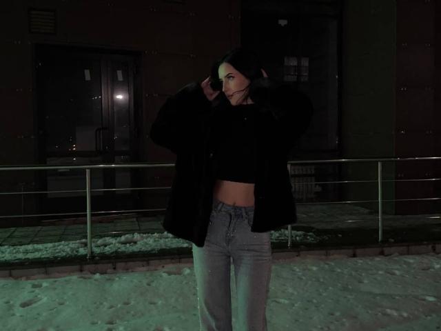BessieSevcik con un cappotto nero, che lascia scoperto il ventre, indossa jeans grigi, è in piedi nella neve di notte, toccandosi i capelli scuri, una luce verde sul viso, un edificio scuro sullo sfondo..