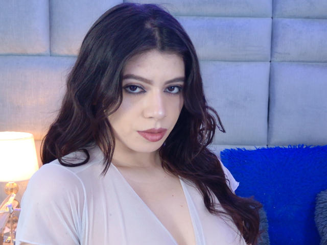 Linda Mancini, avec ses cheveux bruns ondulés, ses yeux bleus et son rouge à lèvres rose, porte un haut blanc transparent qui révèle ses seins, et elle est assise devant une lampe allumée et un coussin bleu..