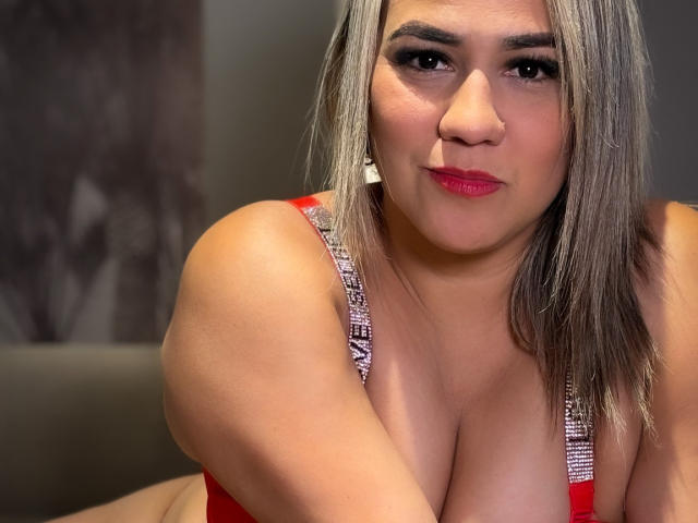 Camila Noer, com pele marrom média, cabelo grisalho, batom vermelho e um sutiã vermelho com alças prateadas, exibindo seus seios grandes e arredondados e um sorriso confiante..