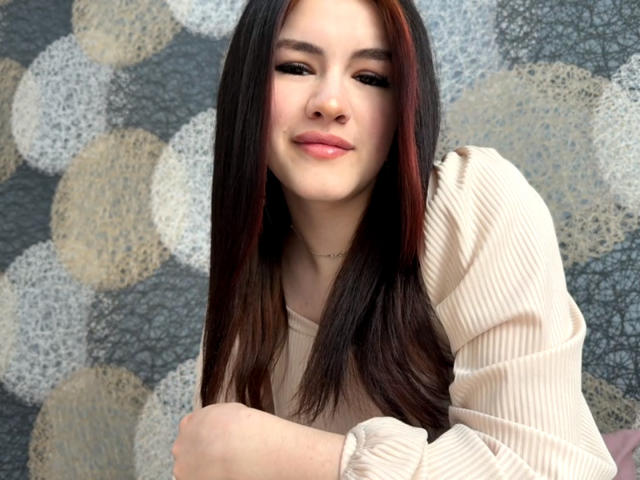 VersieUthe has long, dark brown hair with red highlights, wearing a beige ribbed shirt, sitting with a subtle smile, and a delicate necklace, showing a relaxed, cute look.