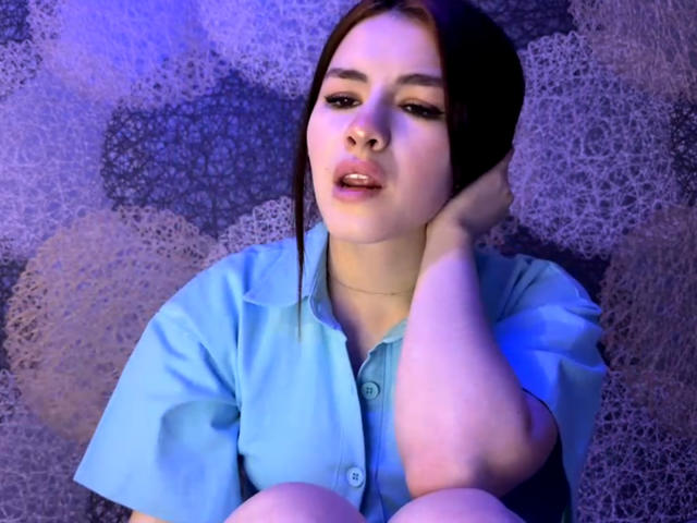VersieUthe has puffy lips, brown hair, and a blue shirt. She's resting her head on her hand, looking slightly annoyed with a slight open-mouth expression.