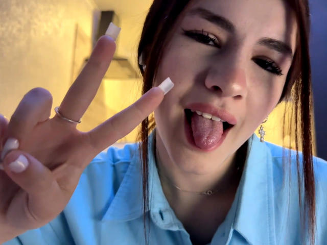 VersieUthe has pale skin, brown hair, and is sticking out her tongue with her pink tongue showing, making a peace sign with her white-manicured fingers. She's wearing a blue shirt.
