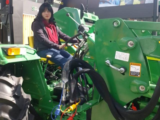IssisDiosa sits on a green tractor with thick black tires, wearing a red shirt and blue jeans, holding the steering wheel, with cables and metal parts visible.