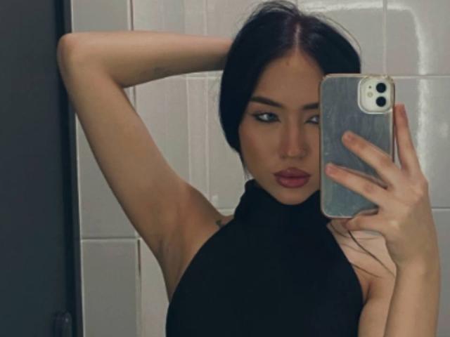DiannLamphear hat dunkles Haar, trägt ein schwarzes Top mit Halter und posiert mit einem Arm erhoben, wobei sie ein Telefon hält und in einem Selfie im Spiegel ihre Brüste zur Schau stellt..