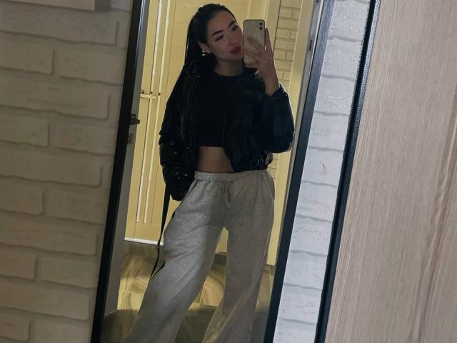 DiannLamphear, avec de longues tresses, portant un crop top noir, une veste noire et un pantalon de jogging gris, prend un selfie devant un miroir, montrant sa silhouette fine et ses fesses discrètes..