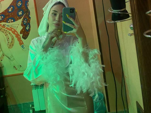 DiannLamphear enveloppée dans une serviette blanche avec des manches duveteuses, tenant un téléphone, autoportrait pris dans un miroir, lumière verte, serviette sur la tête, ongles jaunes, tatouages visibles, sourire doux, pose aguicheuse..