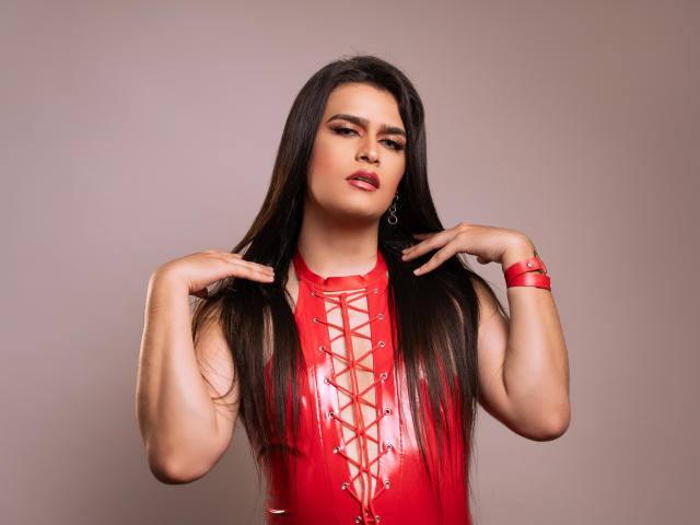 LilithBlackHeart con lunghi capelli scuri, un corsetto in lattice rosso con lacci frontali, un braccialetto rosso, mani vicino alla testa, espressione sicura, rossetto rosa, occhi marroni, pelle abbronzata, sexy, attraente..