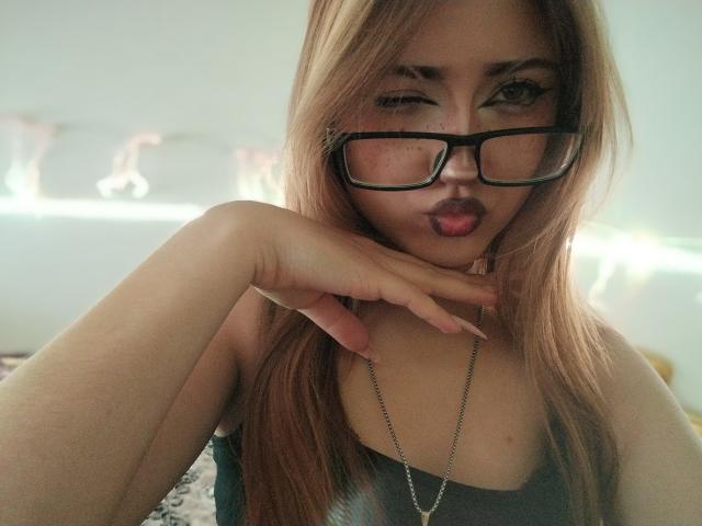 ShainaBallensky with long blonde hair, black glasses, and a necklace, sticking her tongue out, hand near her chin, wearing a gray top, showing her tits slightly.