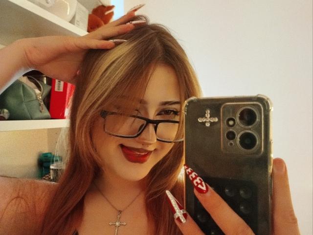 ShainaBallensky with blonde hair, glasses, red lipstick, smiling, holding phone, cross necklace, red nails, hand in hair, sexy, cute, mirror selfie, phone with camera, white wall background.