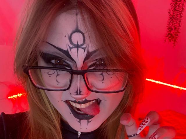 ShainaBallensky with goth makeup, black eyeliner, nose ring, and black lipstick, tongue out, wearing glasses, red light, and showing ring with white dots.